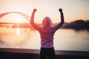 Happy female jogger standing and enjoying in beautiful river sunrise. Winning gesticulation with hands up. View from the back.